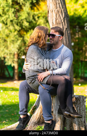 Junge schwangere Frau saß auf ihre Ehemänner schoss, als sie gemeinsam Entspannen im Freien auf dem Baumstumpf in einem Herbst Park Stockfoto