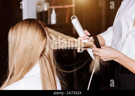 Beauty und Hair Care Konzept. Schöne fröhliche blonde Frau tragen weisse Shirt zu ihrer Freundin Haar styling mit Haar Eisen. Stockfoto