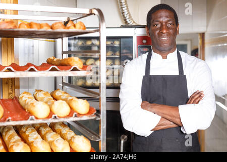 Zuversichtlich Baker in schwarze Schürze mit Waffen in der Nähe von professionellen Brotbackofen in kleine Bäckerei gekreuzt Stockfoto