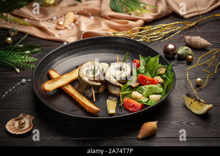 Gebackener Wolfsbarsch mit Auberginen, Mozzarella mit frischem Salat und Grissini Stöcken in einer schwarzen Platte auf einem dunklen Hintergrund Stockfoto