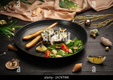 Gebackener Wolfsbarsch mit Auberginen, Mozzarella mit frischem Salat und Grissini Stöcken in einer schwarzen Platte auf einem dunklen Hintergrund Stockfoto