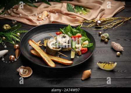 Gebackener Wolfsbarsch mit Auberginen, Mozzarella mit frischem Salat und Grissini Stöcken in einer schwarzen Platte auf einem dunklen Hintergrund Stockfoto