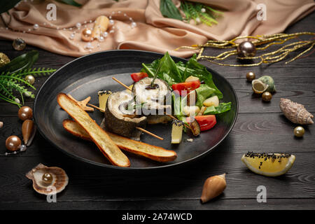 Gebackener Wolfsbarsch mit Auberginen, Mozzarella mit frischem Salat und Grissini Stöcken in einer schwarzen Platte auf einem dunklen Hintergrund Stockfoto