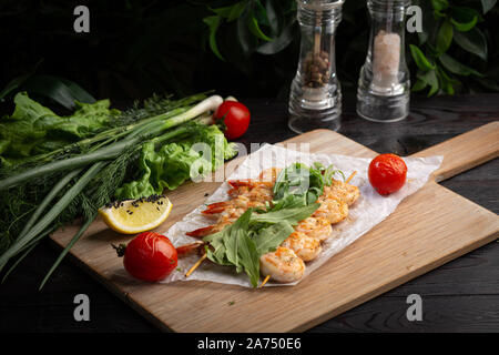 Garnelen Spieße auf einem Spieß mit Rucola, gebackene Tomaten und Zitrone auf einem Bambus Board auf einem dunklen Hintergrund Stockfoto