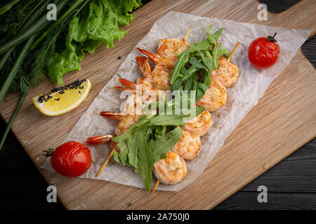 Garnelen Spieße auf einem Spieß mit Rucola, gebackene Tomaten und Zitrone auf einem Bambus Board auf einem dunklen Hintergrund Stockfoto