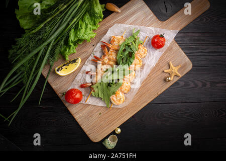 Garnelen Spieße auf einem Spieß mit Rucola, gebackene Tomaten und Zitrone auf einem Bambus Board auf einem dunklen Hintergrund Stockfoto