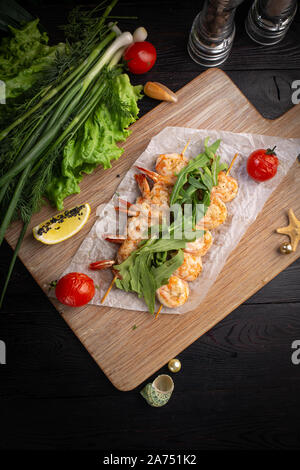 Garnelen Spieße auf einem Spieß mit Rucola, gebackene Tomaten und Zitrone auf einem Bambus Board auf einem dunklen Hintergrund Stockfoto