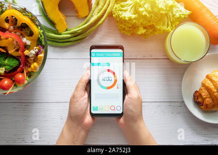 Kalorien zählen und die Lebensmittelüberwachung Konzept. Frau mit Kalorienzähler Anwendung auf Ihrem Smartphone mit Salat, Gemüse, Orangensaft und einem Croissant auf d Stockfoto