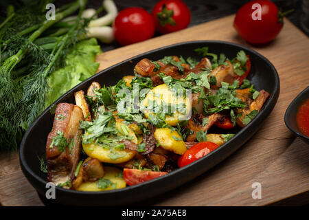 Gebratenes Lamm in einem schwarzen ovalen Schild auf einem Holzbrett auf einem dunklen Hintergrund. Lamm, Kartoffel, rote Zwiebel, Frühlingszwiebel, Kirsche Stockfoto