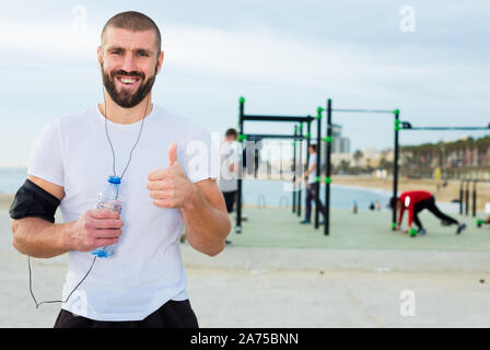 Junge athletischer Mann mit einer Flasche Wasser in den Händen und Kopfhörer steht auf einem offenen Sportplatz Stockfoto