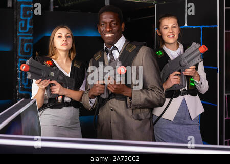 Afrikanische Mann und zwei fröhliche kaukasischen Frauen holding Laser Gewehren und auf der Laser tag Zimmer posing Stockfoto