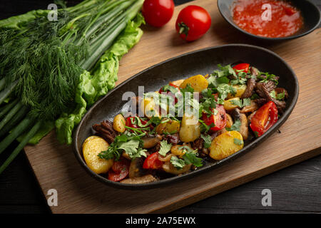 Roastbeef in einem schwarzen ovalen Schild auf einem Holzbrett auf einem dunklen Hintergrund. Lamm, Kartoffel, rote Zwiebel, Frühlingszwiebel, Kirsche Stockfoto