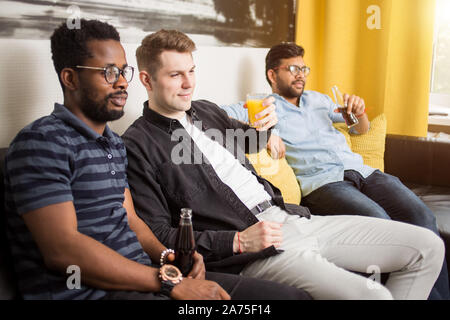 Freundschaft, Sport und Unterhaltung Konzept. Seitenansicht der drei Freunde Fernsehen trinken kohlensäurehaltige Getränke und Saft auf der Couch zu Hause Witz sitzen Stockfoto
