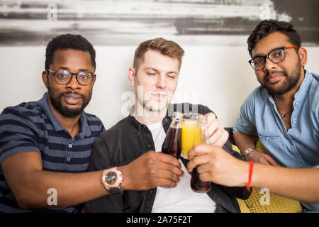Closeup Portrait von drei schönen multi-ethnische männliche Freunde im casual Outfits klirrenden Flaschen Getränke Lächeln auf die Kamera schaut. Menschen, Männer, Leis Stockfoto