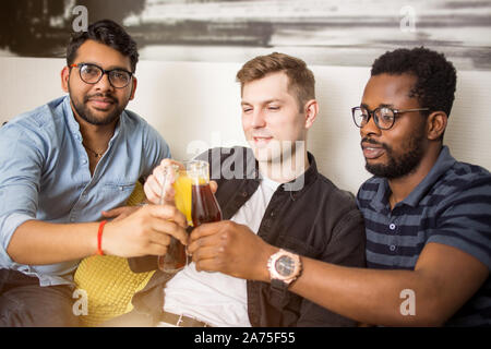 Drei stattliche multirassischen Freunde in legere Kleidung sitzen auf der Couch klirrenden Flaschen Getränke Lächeln auf die Kamera schaut. Fokus auf Getränke. Menschen, Stockfoto