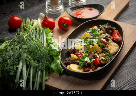 Roastbeef in einem schwarzen ovalen Schild auf einem Holzbrett auf einem dunklen Hintergrund. Lamm, Kartoffel, rote Zwiebel, Frühlingszwiebel, Kirsche Stockfoto