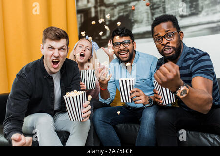 Fröhlich aufgeregt Gruppe der multirassischen Freunde sitzen auf der Couch essen Popcorn, watching Sport Spiel im Fernsehen, schreien, feiern Ziel. Freizeit, Sport, Stockfoto