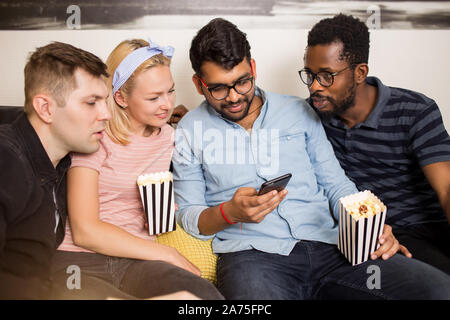 Freundschaft, Technologie Konzept. Vier junge Leute sitzen Zuhause und Handy. Gruppe von Freunden sitzen auf dem Sofa essen Popcorn lächelnd Stockfoto