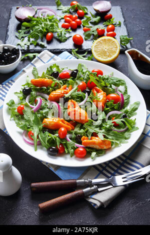 Nahaufnahme der Krabbe schellen Rucola Salat mit schwarzen Oliven, Tomaten und roten Zwiebeln auf einem weißen Teller serviert, auf einer konkreten Tabelle mit Zutaten, Stockfoto