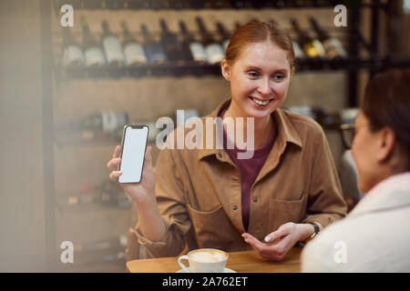 Junge rothaarige Frau mit Mobiltelefon und etwas Übersicht zu ihrer Freundin, während Sie trinken Kaffee im Cafe Stockfoto