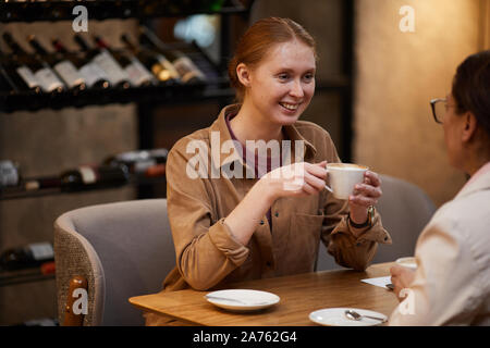 Lächelnde junge Frau hat ein Treffen mit ihrer Freundin im Café sie Kaffee trinken und reden miteinander Stockfoto