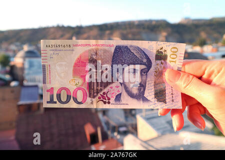 Woman's Hand halten Sie die Vorderseiten der Georgischen 100 Lari Banknote mit verschwommenen Tiflis Luftaufnahme im Hintergrund Stockfoto