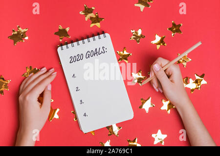 Woman's Hände halten Bleistift in der Nähe der Notepad mit Text 2020 Ziele auf dem roten Hintergrund mit sternförmigen Konfetti. Stockfoto