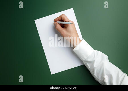 Männliche Hand mit Stift und schreiben auf leeres Blatt auf grünem Hintergrund für Text oder Design. Leere Vorlagen für Kontakt, Werbung oder für den Einsatz in Unternehmen. Finanzen, Büro, Einkäufe. Mock up. Copyspace. Stockfoto