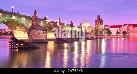 Prag am Sonnenuntergang, panorama bild. Beleuchtete Charles Bridge, Bridge Tower und alte Gebäude am Flussufer unter purple sky mit goldenen Reflexion Stockfoto