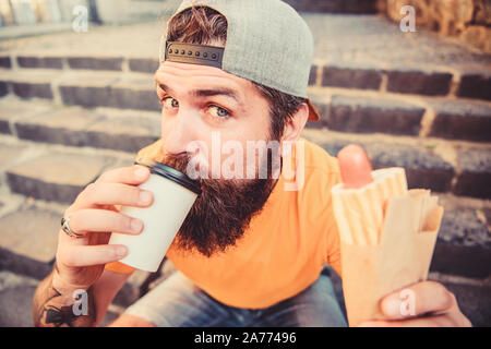 Trinken Sie etwas anders. Kaukasische hipster Trinken Essen trinken mit Hot Dog. Man nimmt einen Schluck frischen Kaffee trinken im verfügbaren cup Bärtigen. Ernährung Essen und Trinken. Stockfoto