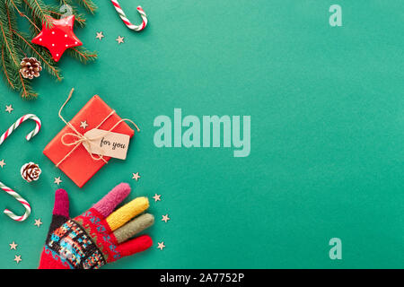 Kreative Weihnachten flach Hintergrund in grün und rot auf grünem Papier mit copy-Platz. Hand in reich verzierten wolle Handschuh, Zuckerstangen, verpackte Geschenk mit Karte Stockfoto