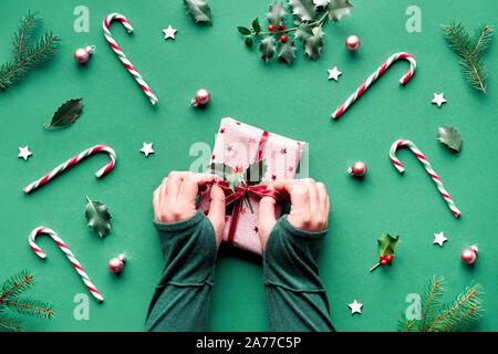 Trendy Weihnachten flach auf grünem Papier Hintergrund mit Zuckerstangen, Holly und Tannen Zweigen, Sterne aus Holz und Glas Trinkets. Weibliche Hände in Grün shir Stockfoto