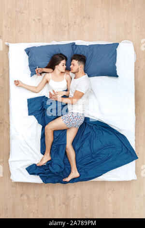 Bedtime. Junger Mann surfen Smartphone nachdenklich, Bein auf Sleeping Woman hugging schlafend auf dem Bett Top View Stockfoto