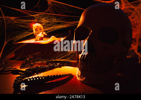 Menschlicher Schädel im orangen Kerzenlicht, umgeben von Spinnweben, auf dem Tisch mit altem Buch, Edelstein und Anhänger Stockfoto