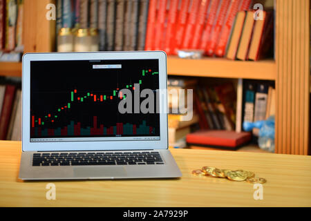 Auf dem Tisch golden Bitcoins und Laptop mit Börse Grafik auf dem Bildschirm, verschwommenes Regal mit Büchern in der background-image Stockfoto