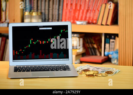 Auf dem Tisch golden Bitcoins und Laptop mit Börse Grafik auf dem Bildschirm, verschwommenes Regal mit Büchern in der background-image Stockfoto