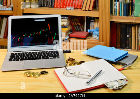 Auf dem Tisch golden Bitcoins, Notebook, fernglaeser und Laptop mit Börse Grafik auf dem Bildschirm, verschwommenes Regal mit Büchern in der background-image Stockfoto
