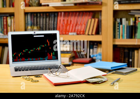 Auf dem Tisch golden Bitcoins, Notebook, fernglaeser und Laptop mit Börse Grafik auf dem Bildschirm, verschwommenes Regal mit Büchern in der background-image Stockfoto