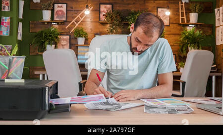 Designer Junge, der am Schreibtisch sitzt und macht einige Anmerkungen zu einigen gedruckten Blätter. Loft Büro Stockfoto