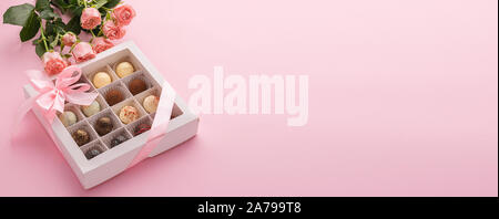Eine Box mit verschiedenen Schokoladen ist mit einem rosa satin schleife Blumen von einer Rose auf einem rosa Hintergrund eingerichtet. Blick von oben. Kopieren Sie Platz. Horizontale Stockfoto