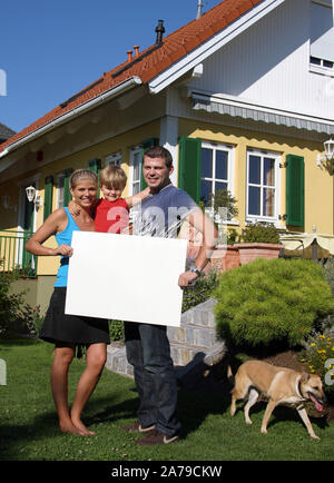 Junge Familie mit Art vor neugekauftem Haus, hält weisses Schild, frei für beliebigen Text, 30,35, 5, Jahre, HERR: Ja Stockfoto