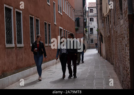 Zwei junge Damen mit einem Kerl entlang eines gepflasterten sonnenbeschienenen Gasse in Venedig suchen glücklich und sorgenfrei während des Chats. Stockfoto