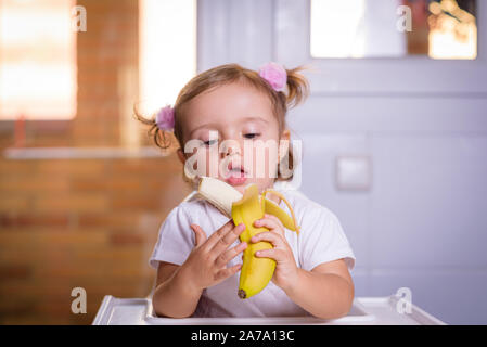 Cute Baby 1,4 Jahre alt auf hohen Kinderstuhl sitzen und essen eine Banane allein in Weiß Küche Stockfoto