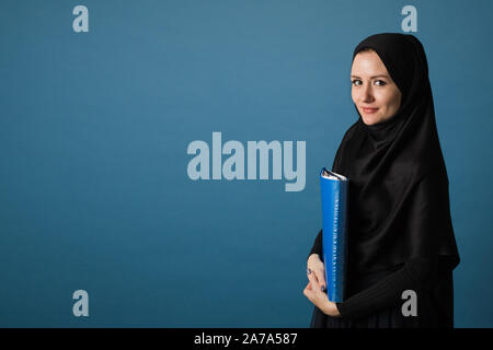 Schönen muslimischen Mädchen Sekretärin oder Office Manager im hijab steht mit einem Ordner von Papieren, die in ihre Hände auf einem blauen Hintergrund im Studio. Konzept der Stockfoto