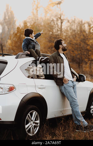 Gerne Vater stanting in der Nähe des Auto mit seinem kleinen Sohn sitzen auf Auto und zeigen auf Sky. Stockfoto