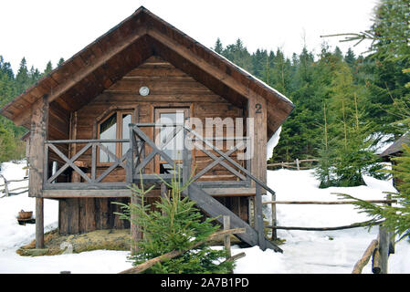 Holz-, braunes Haus auf dem Berg, unter dem grünen Nadelwald. Winter Natur, bewölkter Tag, eine Menge weißer Schnee. Stelzenhaus mit hölzernen stairca Stockfoto