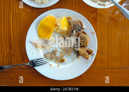 Auf einer weißen Platte sind die Knochen eines gegessen gebratener Fisch und Zitronenscheiben Haut. Auf braunem Holz- Tabelle ist eine weiße Platte mit einer Gabel und Essensreste Stockfoto