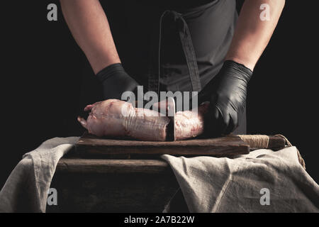 Koch in Schwarzem latex Chunks in Stücke schneidet rohes Fleisch auf einem braunen Holzbrett, kochen auf einem dunklen Hintergrund Stockfoto