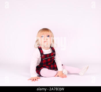 Kleine blonde Kleinkind Mädchen mit großen blauen Augen im roten Kleid auf dem Boden sitzend, weisser Hintergrund. Porträt, Nahaufnahme isoliert Stockfoto