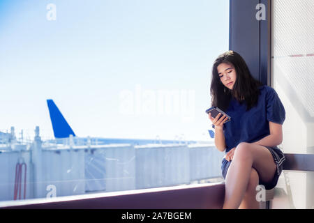 Biracial asiatischen Kaukasischen jugendlich Mädchen sitzt neben sonnigen Fenster am Flughafen, während Ihr Smartphone suchen Stockfoto